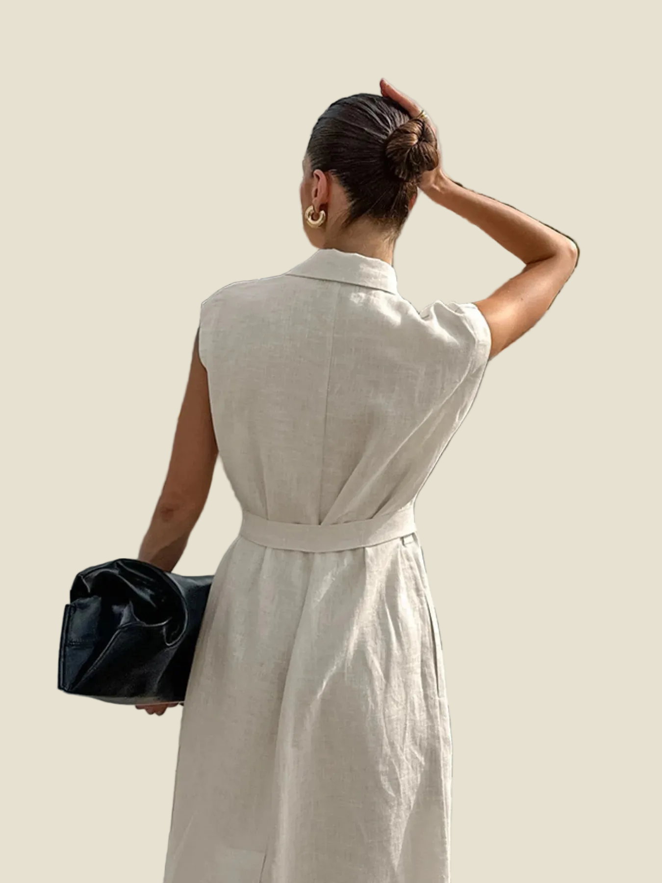 A woman shown from behind wearing a light beige linen belted dress, holding a black clutch bag against a solid beige background.