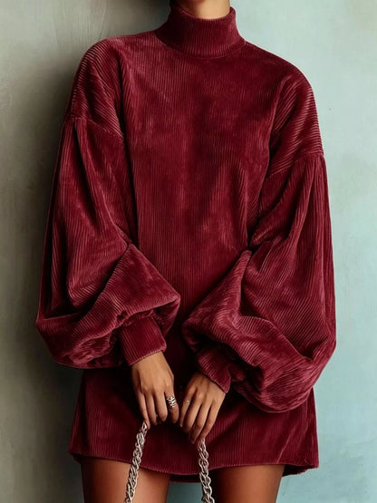 A person wearing an oversized corduroy mock-neck tunic dress in deep burgundy, featuring voluminous balloon sleeves and holding a silver chain handbag against a textured light blue wall.