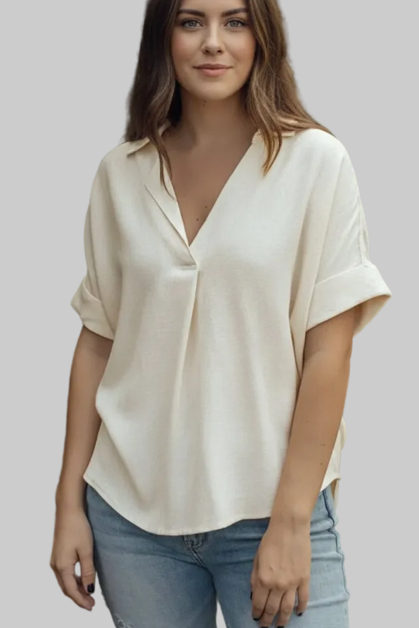 A woman wearing a cream-colored short-sleeve blouse with a V-neckline and rolled cuffs, paired with light blue jeans, set against a solid gray background.