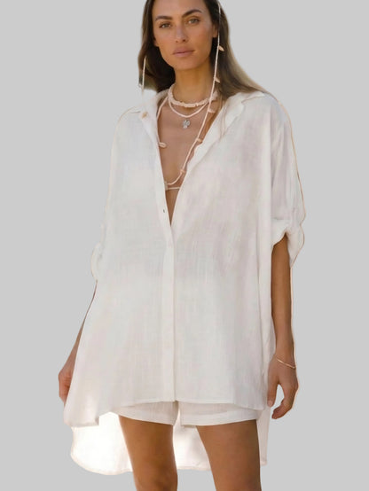 A woman modeling a loose-fitting, oversized white linen button-up shirt dress with short sleeves and a high-low hem, styled with layered pearl necklaces against a solid gray background.