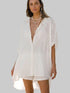 A woman modeling a loose-fitting, oversized white linen button-up shirt dress with short sleeves and a high-low hem, styled with layered pearl necklaces against a solid gray background.
