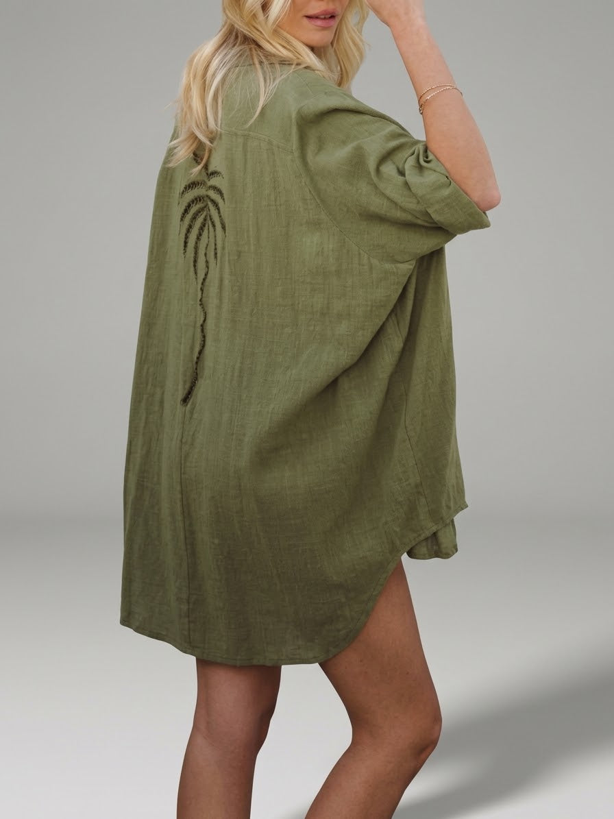 A woman wearing an olive green linen kimono-style cover-up dress with a dark palm tree embroidery on the back, shown from a three-quarters back view against a neutral gray studio background.