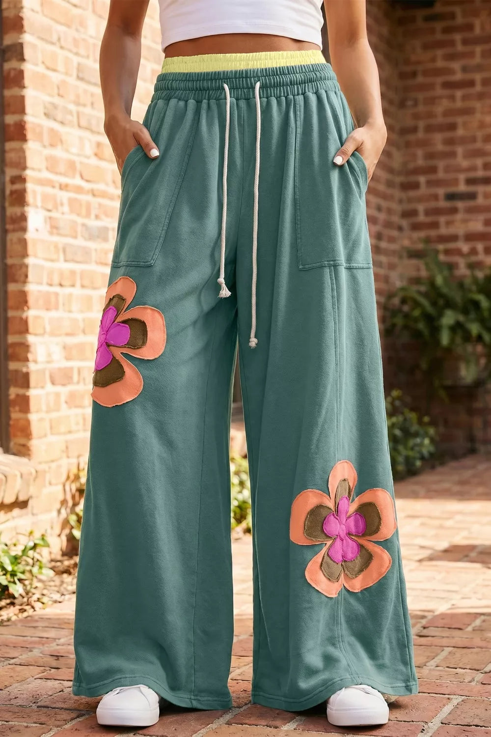 Person wearing teal pants with floral patches in front of a brick wall.