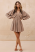 A woman models a taupe-brown mini dress with voluminous balloon sleeves, a smocked bodice, and a V-neckline, standing against a textured beige plaster wall.