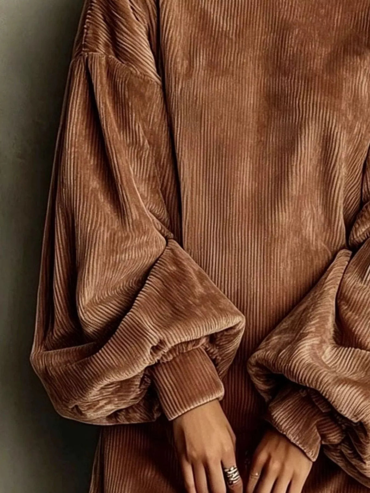A close-up of a person wearing a brown corduroy long-sleeve top or dress with voluminous balloon sleeves and ribbed cuffs, shown against a dark neutral background.