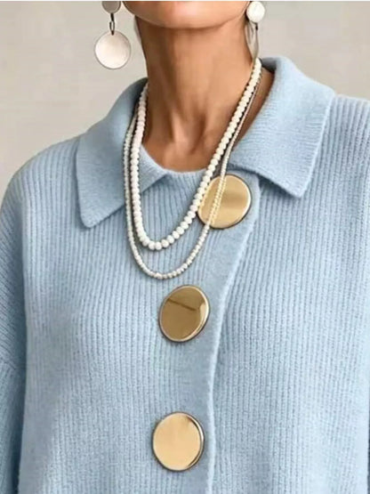 A close-up of a person wearing a light blue knit collared sweater with large round gold buttons, layered with white pearl necklaces and silver drop earrings.