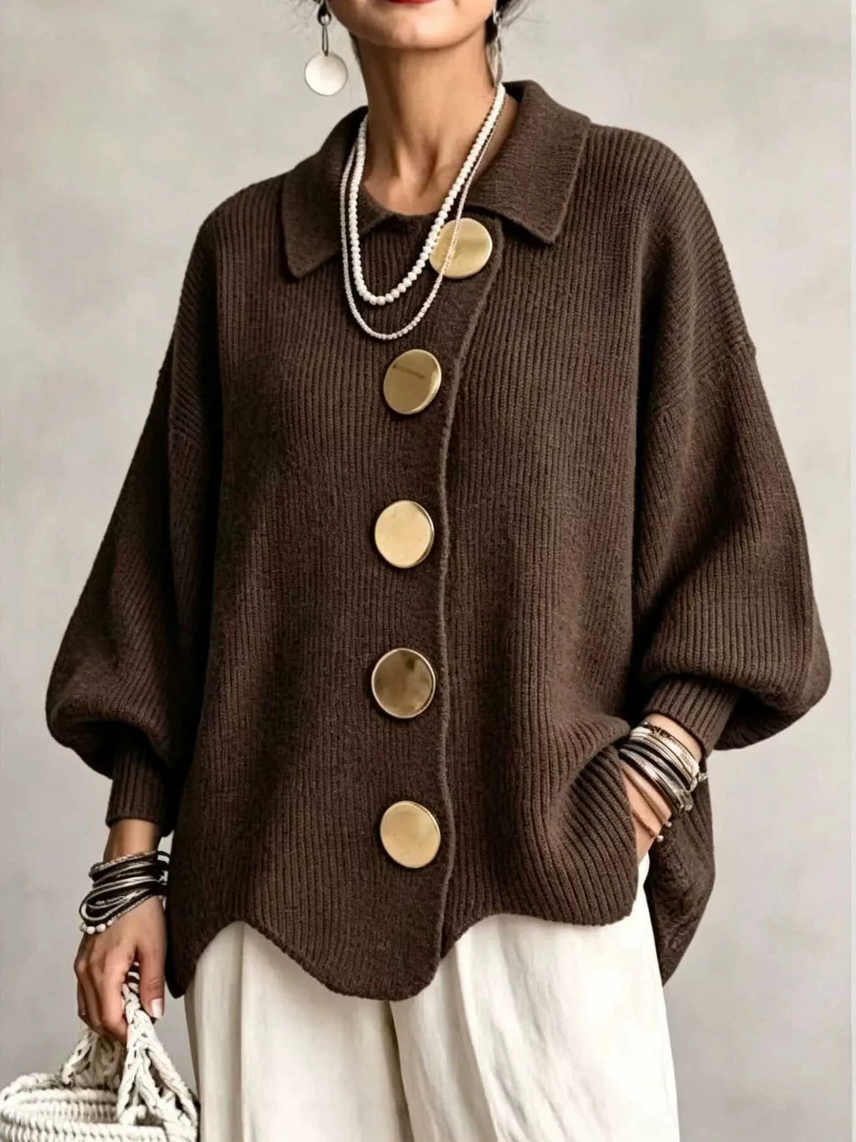 A woman wearing a dark brown ribbed knit cardigan sweater with oversized round gold-tone buttons, paired with white pants and layered pearl necklaces, against a neutral gray background.