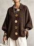 A woman wearing a dark brown ribbed knit cardigan sweater with oversized round gold-tone buttons, paired with white pants and layered pearl necklaces, against a neutral gray background.