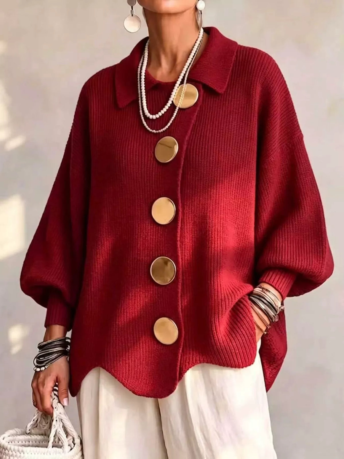 A woman wearing a deep red ribbed knit cardigan with oversized gold buttons, styled with white pants, layered pearl necklaces, and bracelets.