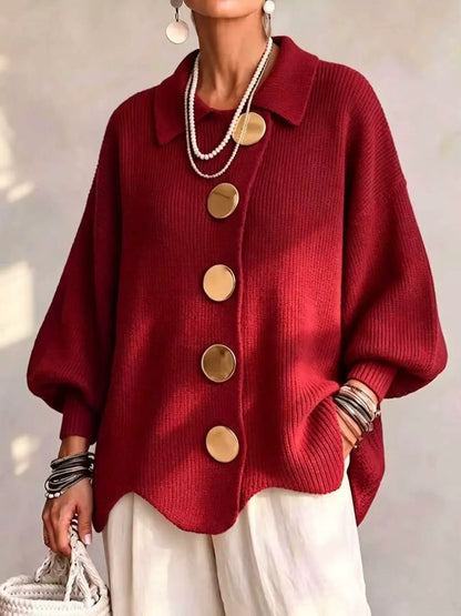 A woman wearing a deep red ribbed knit cardigan with oversized gold buttons, styled with white pants, layered pearl necklaces, and bracelets.