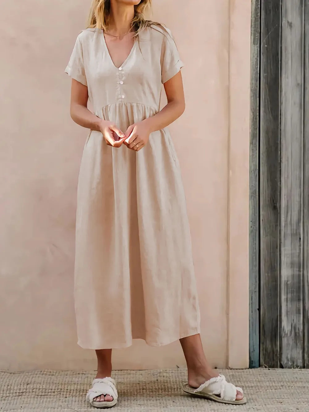 Woman wearing a beige dress is standing against a pink wall.