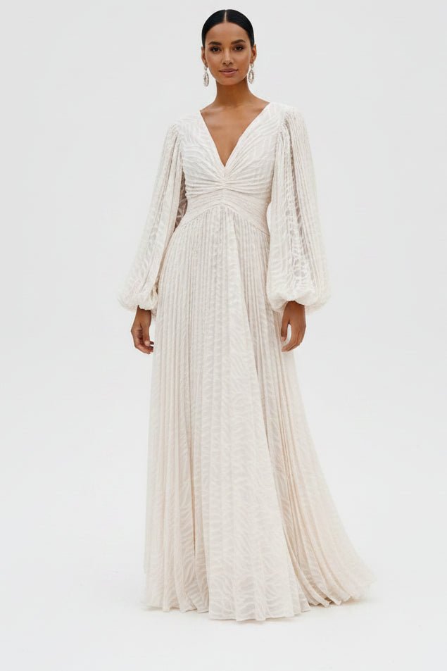 A model wearing a floor-length cream lace gown with long balloon sleeves, a deep V-neckline, and a gathered bodice, photographed against a seamless white studio background.