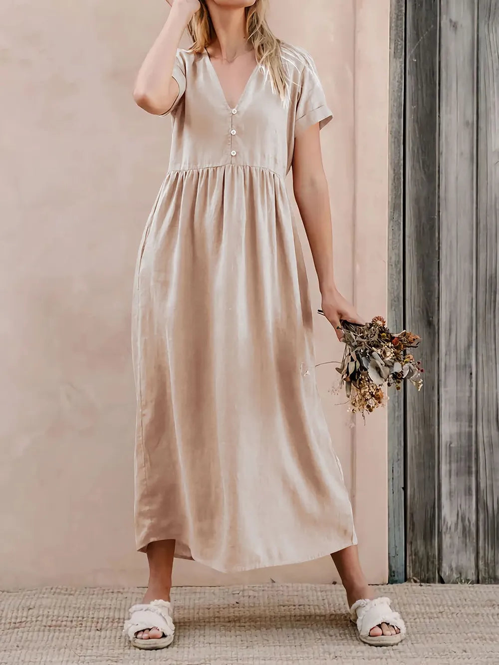 A woman in a beige dress holds dried flowers against a wooden wall.