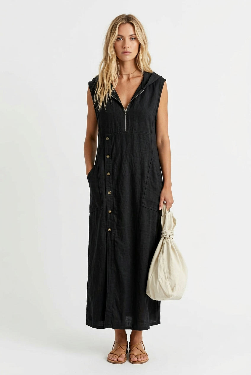 A woman wearing a sleeveless black midi dress with a front zipper, button-down front, and side pockets, holding a cream-colored fabric bag in an outdoor setting with a white picket fence and greenery.