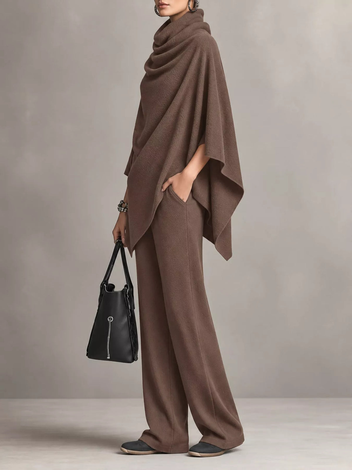 A model wearing a matching brown draped poncho and wide-leg pants set, holding a black handbag against a neutral gray studio background.