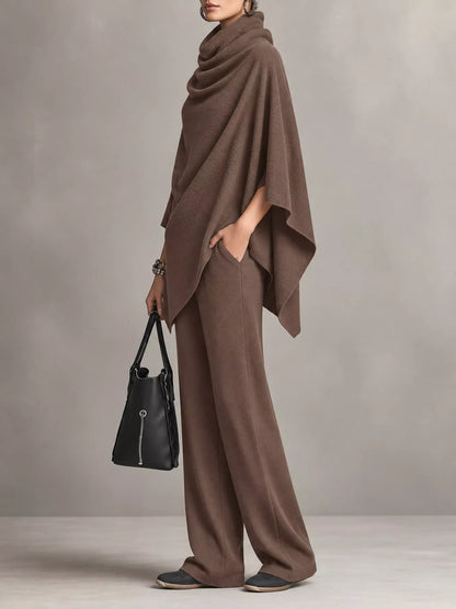 A model wearing a matching brown draped poncho and wide-leg pants set, holding a black handbag against a neutral gray studio background.
