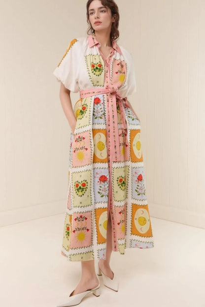 A woman models a long white shirt dress with a colorful patchwork print featuring floral and egg motifs, styled with a pink waist tie and white pointed-toe heels against a cream-colored studio background.