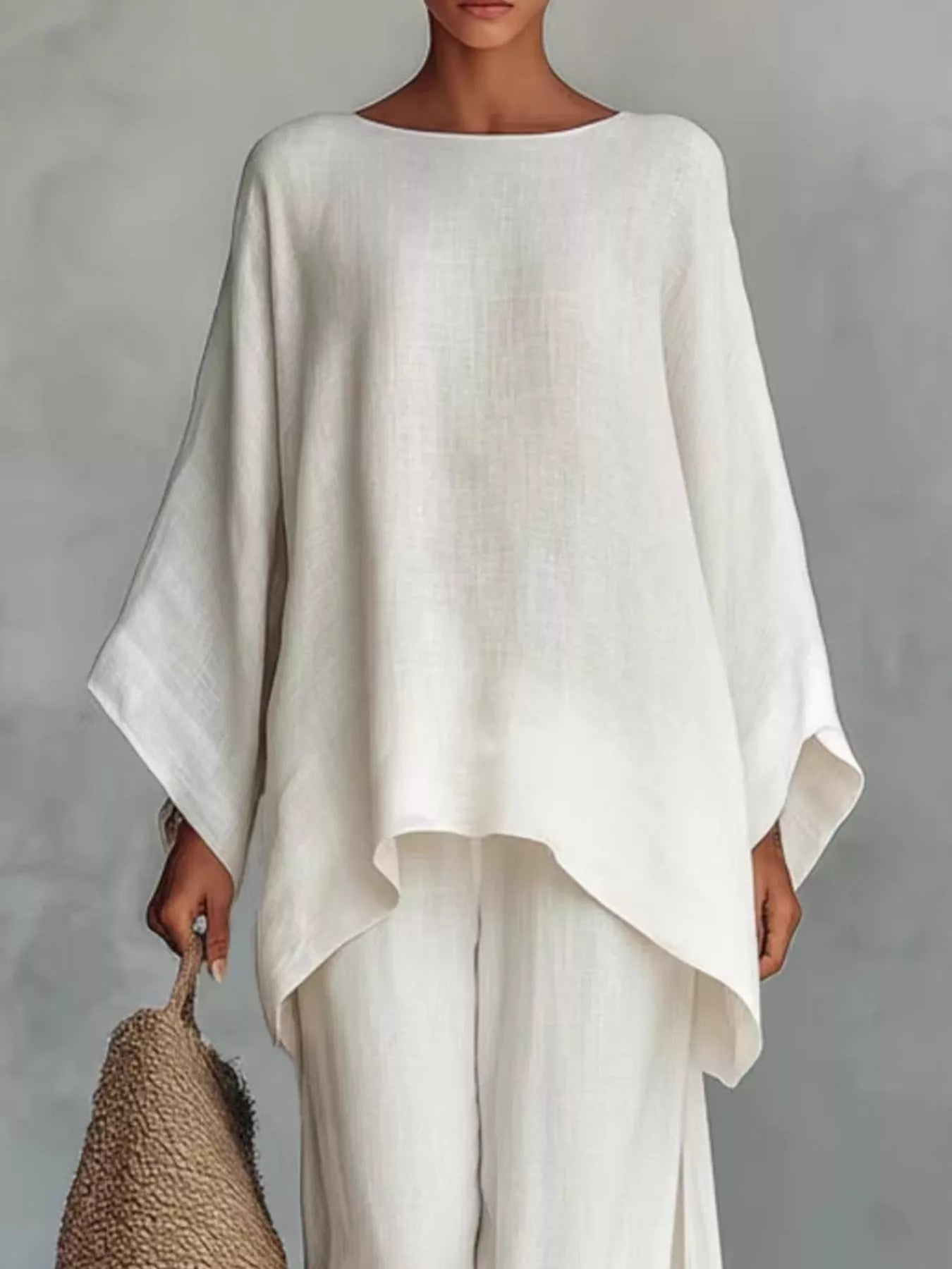 A woman wearing a loose, oversized off-white linen top with wide draped sleeves and matching wide-leg pants, holding a woven straw bag against a neutral gray background.