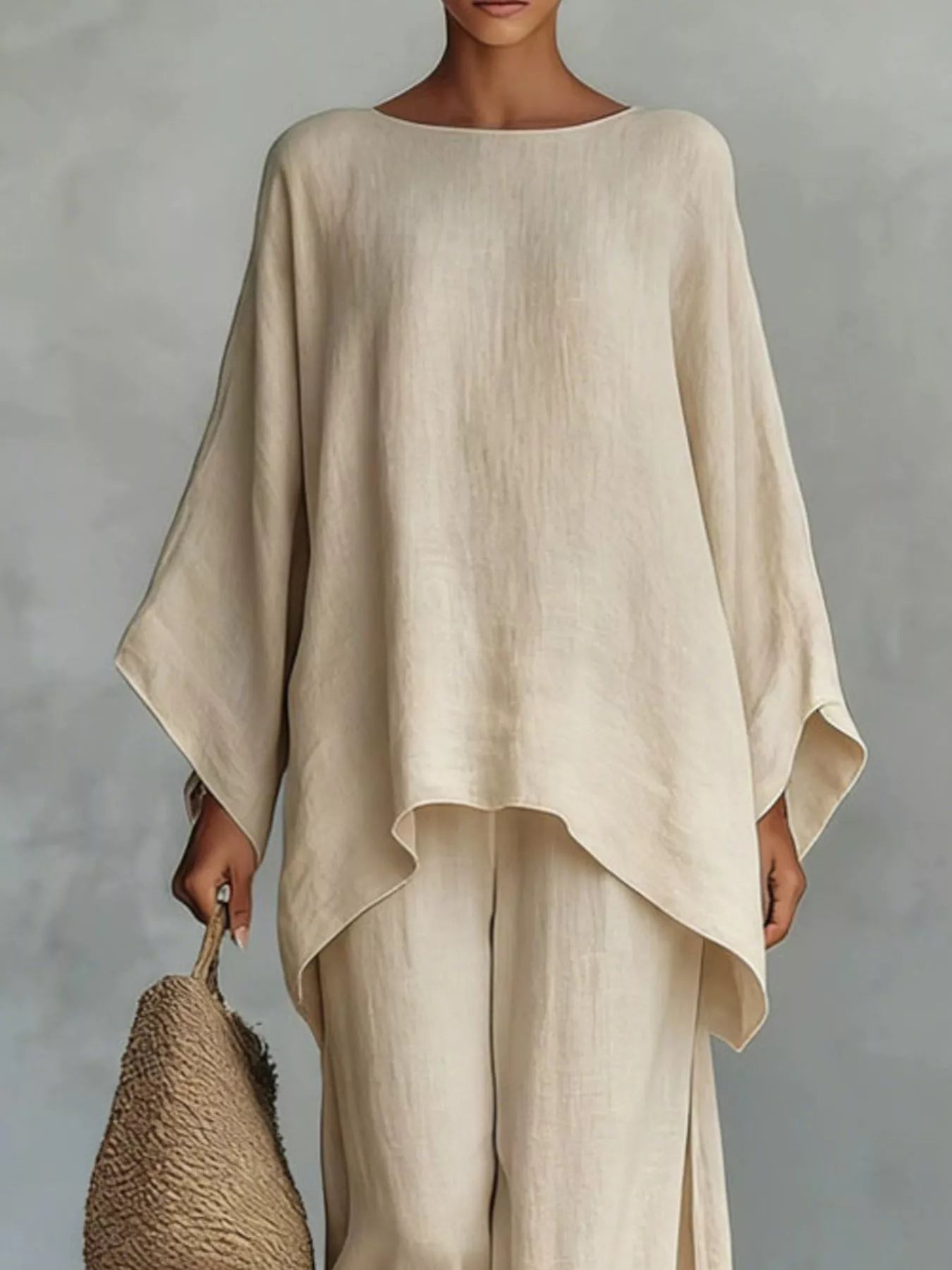 A woman wearing a loose, high-low beige linen top and matching wide-leg pants, holding a woven straw bag against a textured gray studio background.