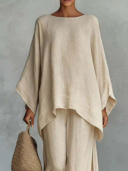 A woman wearing a loose, high-low beige linen top and matching wide-leg pants, holding a woven straw bag against a textured gray studio background.