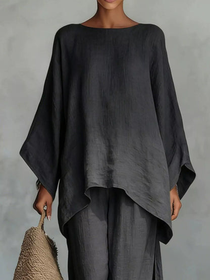 A model wearing a dark charcoal gray linen-blend top with wide draped sleeves and matching wide-leg pants, holding a woven straw bag against a textured gray studio background.