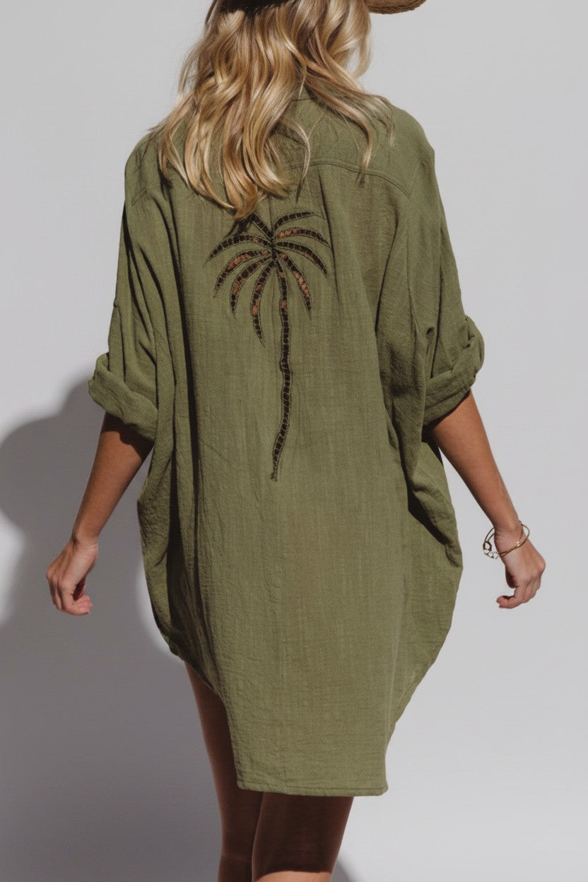 A woman shown from the back wearing an olive green linen shirt dress with a large embroidered palm tree design on the back, set against a plain light gray studio background.