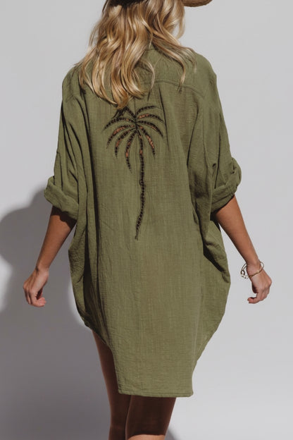A woman shown from the back wearing an olive green linen shirt dress with a large embroidered palm tree design on the back, set against a plain light gray studio background.