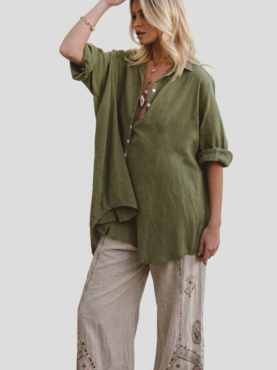 A woman modeling an olive green linen button-up shirt with rolled sleeves and beige patterned linen pants against a solid light gray background.