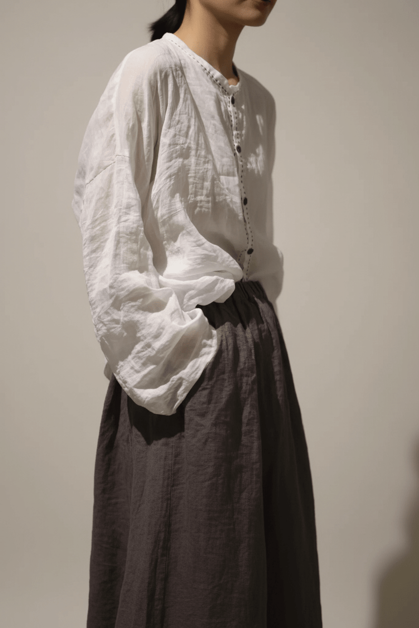 A person wearing a loose white long-sleeve blouse with gathered cuffs and a button placket, paired with a dark skirt, is photographed in a dim indoor setting.