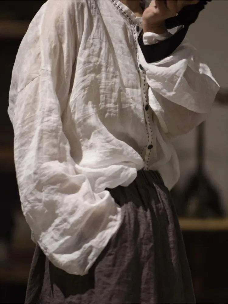 A person wearing a loose white long-sleeve blouse with gathered cuffs and a button placket, paired with a dark skirt, is photographed in a dim indoor setting.