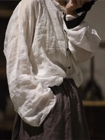 A person wearing a loose white long-sleeve blouse with gathered cuffs and a button placket, paired with a dark skirt, is photographed in a dim indoor setting.