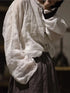 A person wearing a loose white long-sleeve blouse with gathered cuffs and a button placket, paired with a dark skirt, is photographed in a dim indoor setting.
