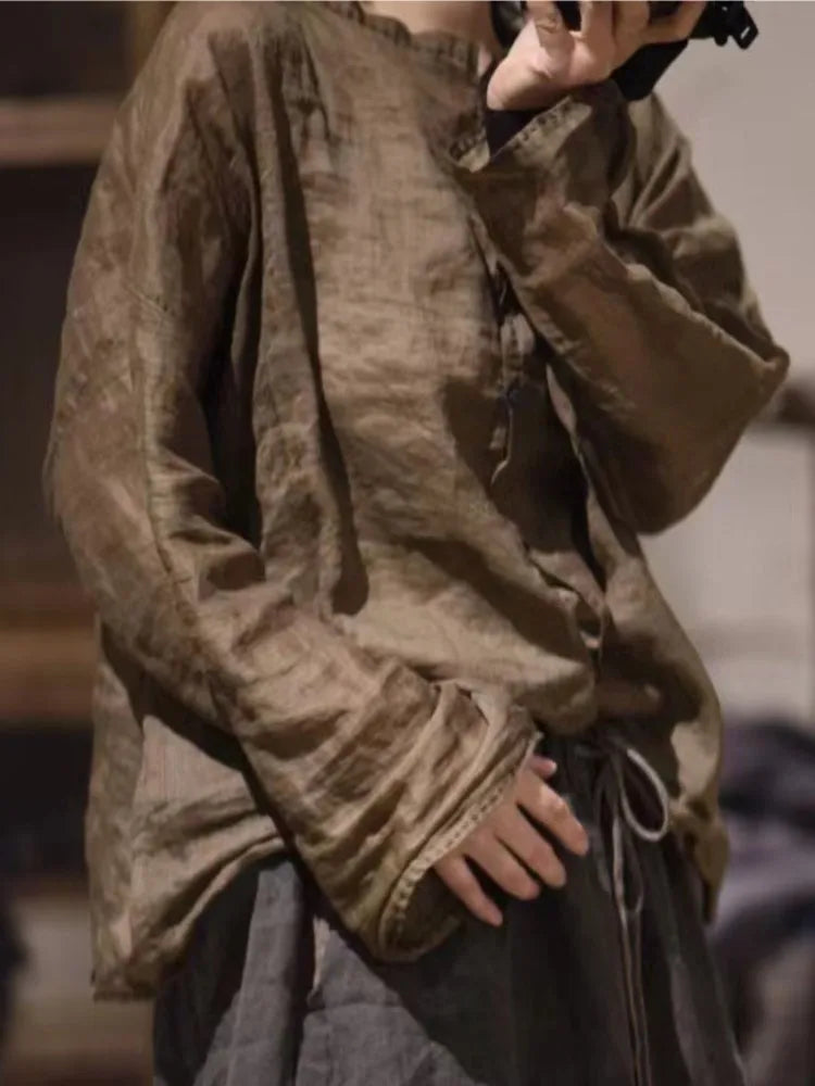 A person wearing a loose, long-sleeve brown linen-like top with a drawstring waist and dark pants, photographed in a dim indoor setting.