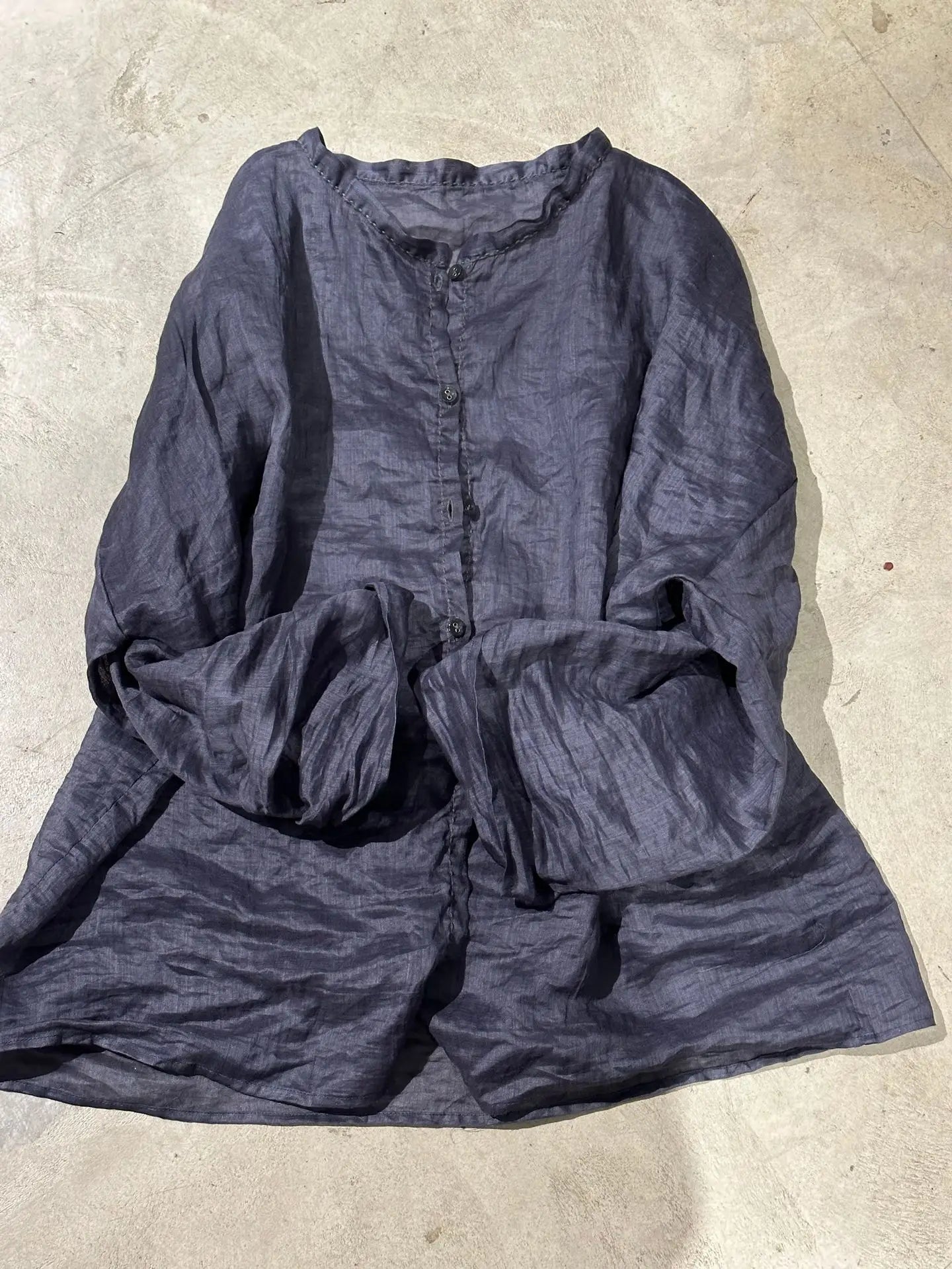 A dark charcoal gray crinkled linen blouse with a buttoned front and ruffled neckline, laid flat on a textured concrete floor.