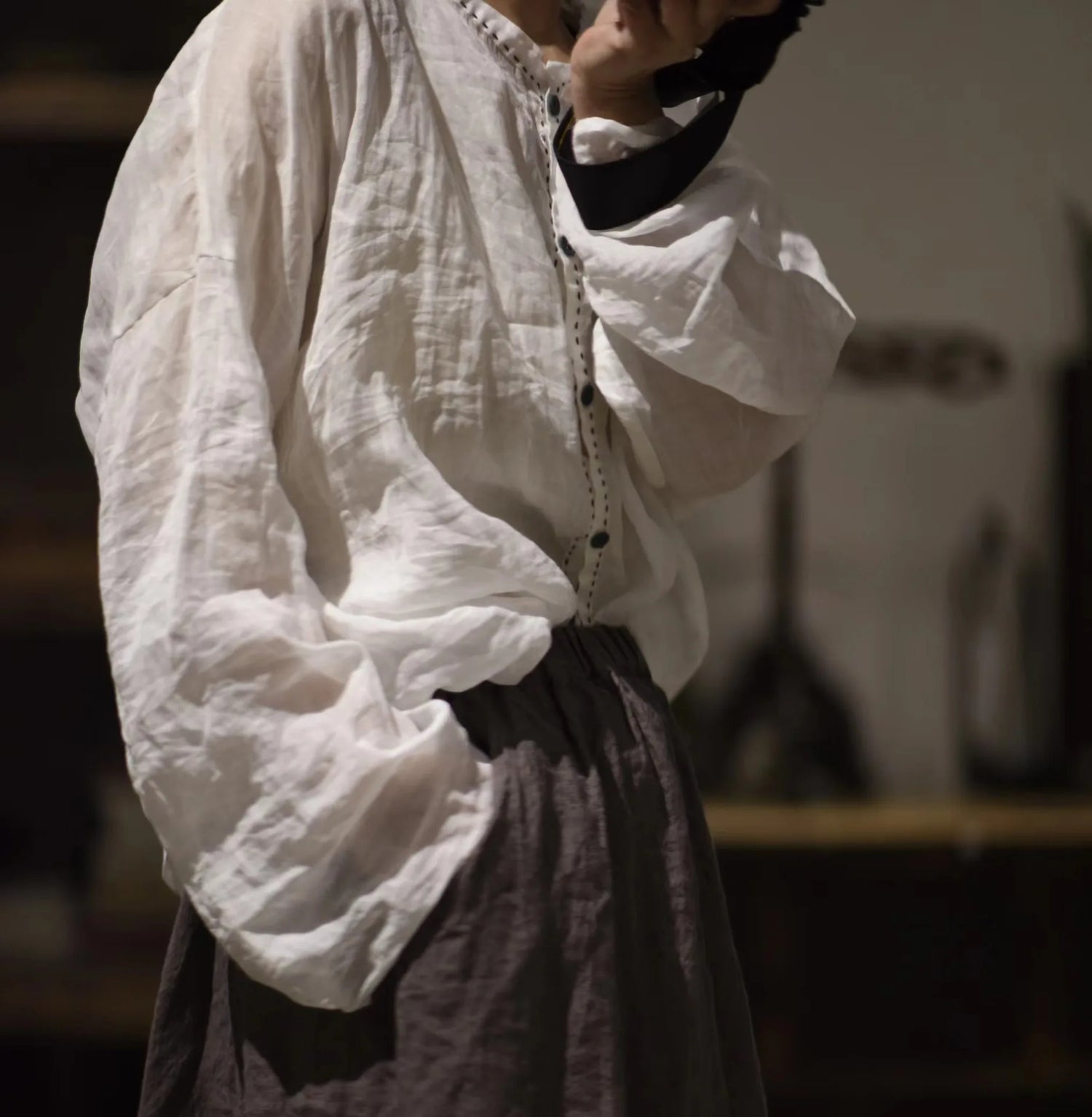A person wearing a loose white long-sleeve blouse with gathered cuffs and a dark brown skirt, photographed in a dim indoor setting with shallow depth of field.
