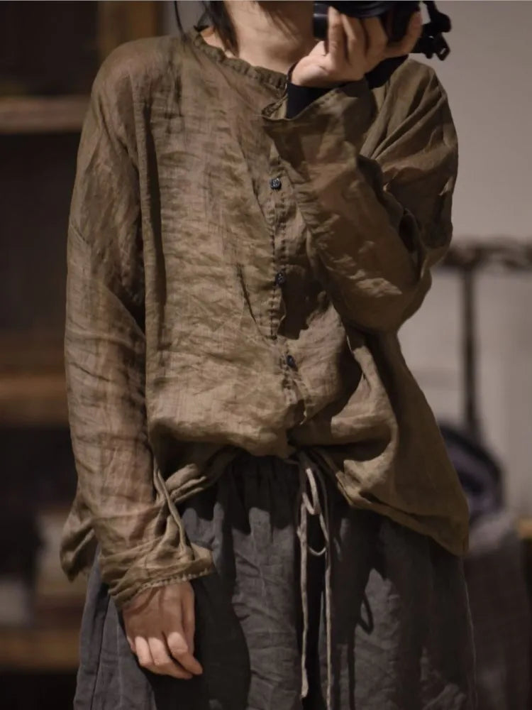 A person wearing a loose, long-sleeve button-front top in a muted brownish-gray linen-like fabric, paired with dark drawstring pants, photographed indoors with a blurred background.