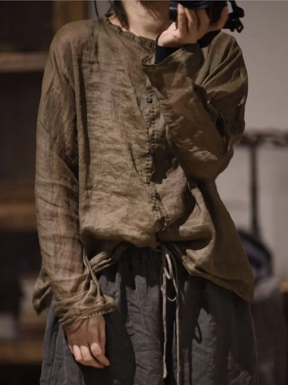 A person wearing a loose, long-sleeve button-front top in a muted brownish-gray linen-like fabric, paired with dark drawstring pants, photographed indoors with a blurred background.