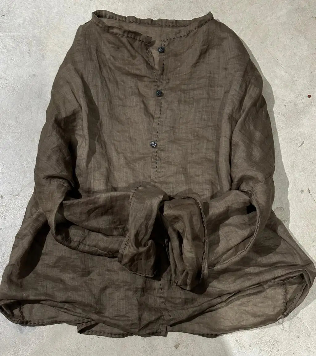 A dark brown, textured linen long-sleeve shirt with a buttoned yoke and a tie detail at the waist, laid flat on a light gray concrete surface.