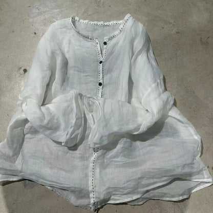 A white long-sleeve linen tunic top with black buttons and decorative stitching, laid flat on a concrete floor.