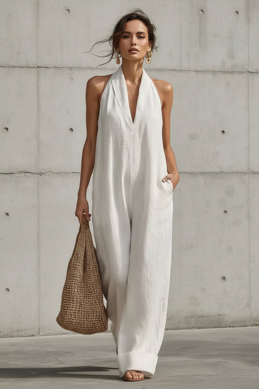 A woman models a white sleeveless halter jumpsuit with wide legs, holding a woven straw tote bag against a concrete wall background.