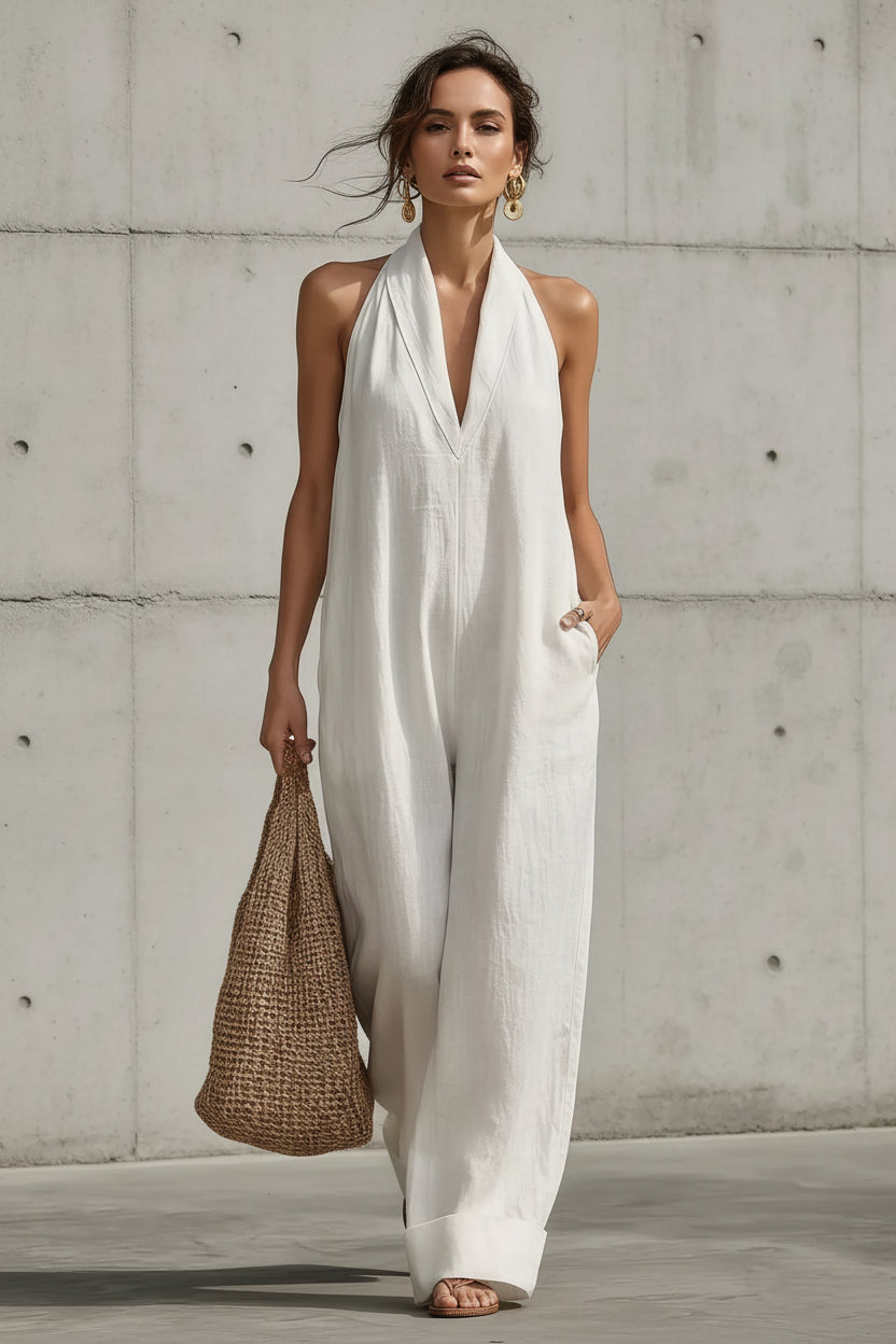 A woman wearing a white sleeveless halter jumpsuit with wide legs, holding a woven straw tote bag against a concrete wall background.