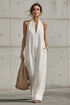 A woman wearing a white sleeveless halter jumpsuit with wide legs, holding a woven straw tote bag against a concrete wall background.