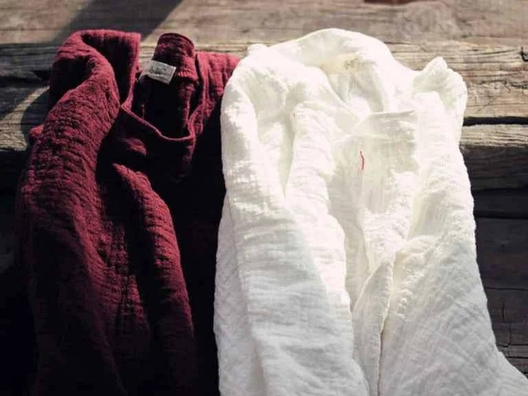 Two textured cotton gauze garments, one in deep burgundy and one in white, draped on a rustic wooden surface.