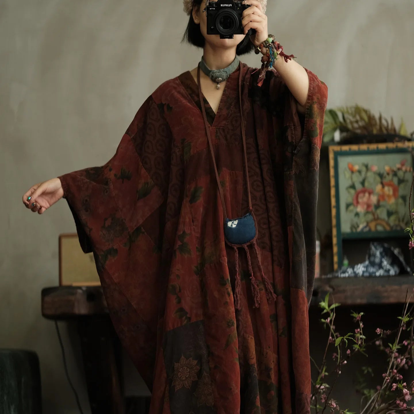 Person wearing a dark red patterned dress with a camera, standing in a room with a decorative background.