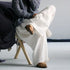 A person seated on a small wooden stool wearing loose white wide-leg pants, a dark blue-gray knit top, and brown slip-on shoes against a plain light gray studio background.