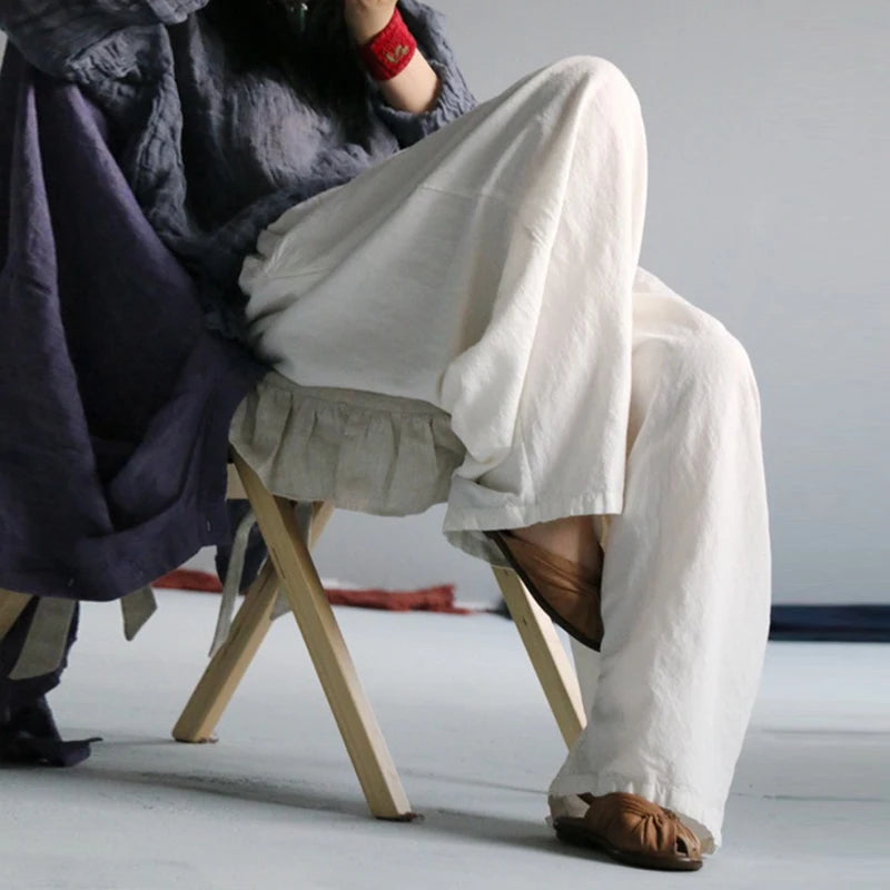 A person seated on a small wooden stool wearing loose, flowing off-white linen pants and brown sandals, with a dark draped garment and red wristband visible against a plain light gray studio background.