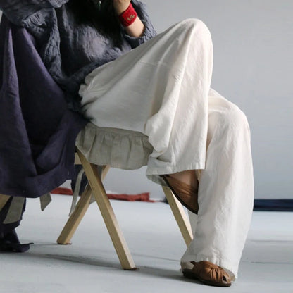 A person seated on a small wooden stool wearing loose, flowing off-white linen pants and brown sandals, with a dark draped garment and red wristband visible against a plain light gray studio background.