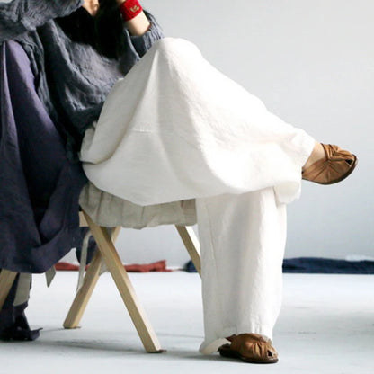 A person seated on a wooden folding chair wearing loose, wide-leg white linen pants and brown leather slide sandals, with a dark textured top partially visible. The photo is cropped to show the lower body and feet against a plain light gray studio background.