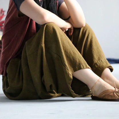 A person sitting on a light gray floor wearing loose, draped olive-brown pants, a dark top, and brown flat shoes.