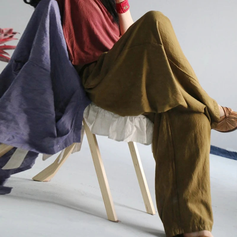 A person seated on a wooden chair wearing loose olive-brown pants and a red top, with a large folded purple textile draped over the left side of the chair in a studio setting.
